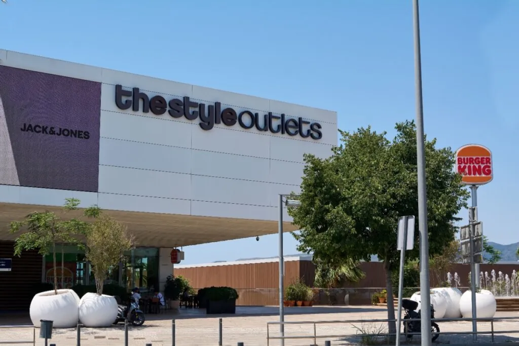 Fachada do espaço The Style Outlets em Barcelona, com logotipo visível e árvores na entrada em um dia ensolarado