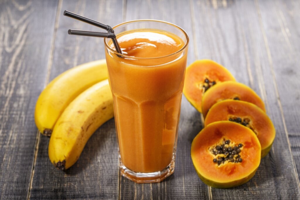 copo de vidro com vitamina laranja e dois canudos de metal. duas bananas e rodelas de mamão com sementes em volta do copo
