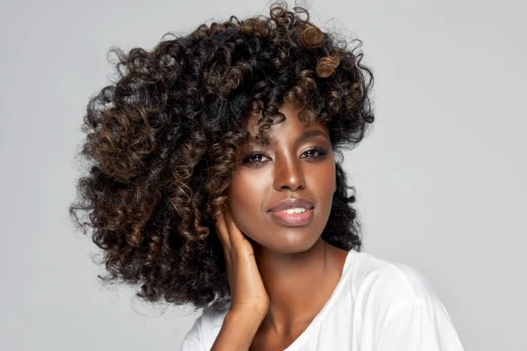 Mulher com cabelo marrom café usando uma blusa branca e posando para foto em frente a um fundo cinza