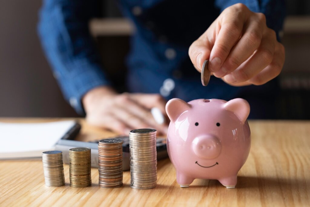 Mão colocando moeda em um cofrinho no formato de porco. Ao lado, moedas empilhadas