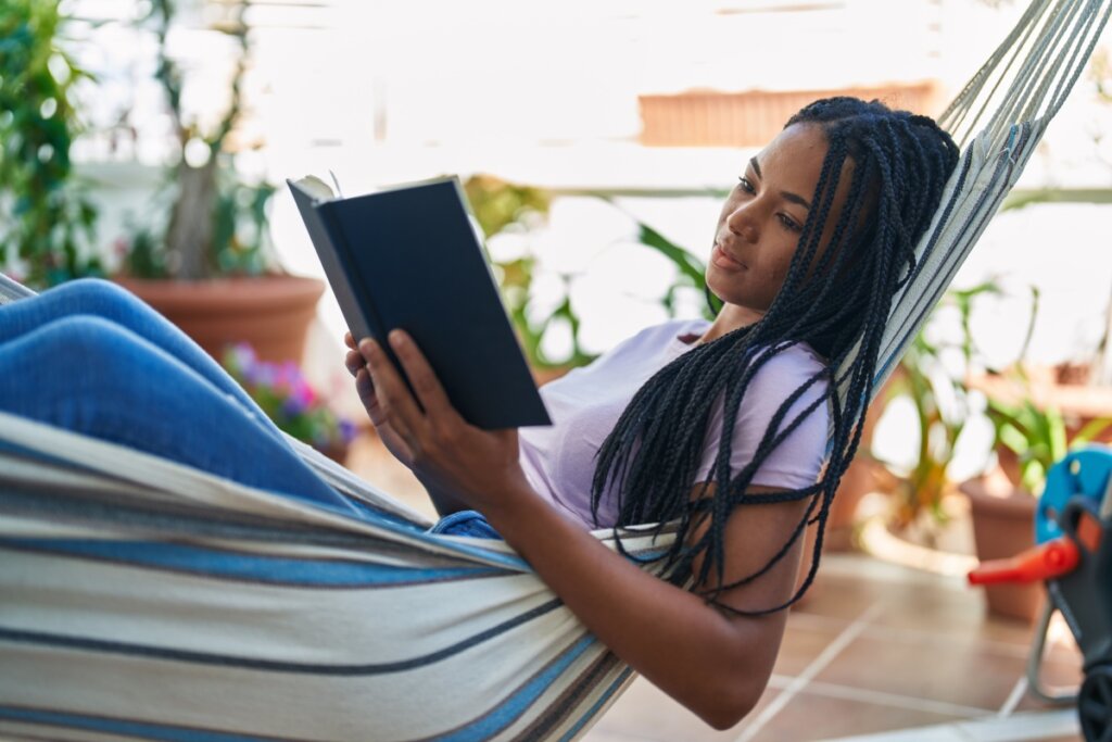 Jovem com o cabelo longo, trançado, usando uma camiseta roxa deitada em rede listrada no quintal com um livro na mão