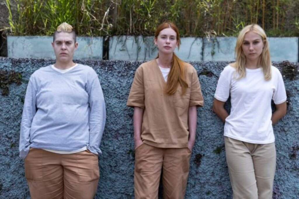 Atrizes Letícia Rodrigues, Marina Ruy Barbosa e Carol Garcia paradas lado a lado e vestindo roupas de presidiárias