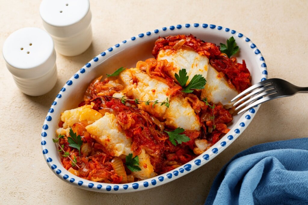 Travessa oval contendo bacalhau ao molho de tomate com cebola, cenoura e ervas