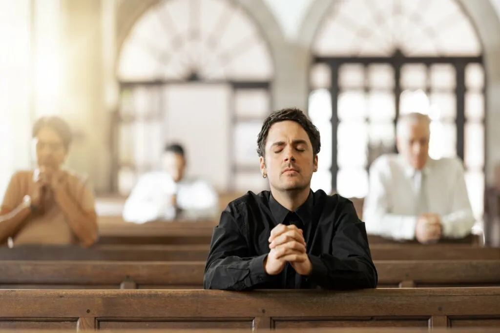 Homem vestindo roupa preta sentado em uma igreja e orando com as mãos juntas e os olhos fechados