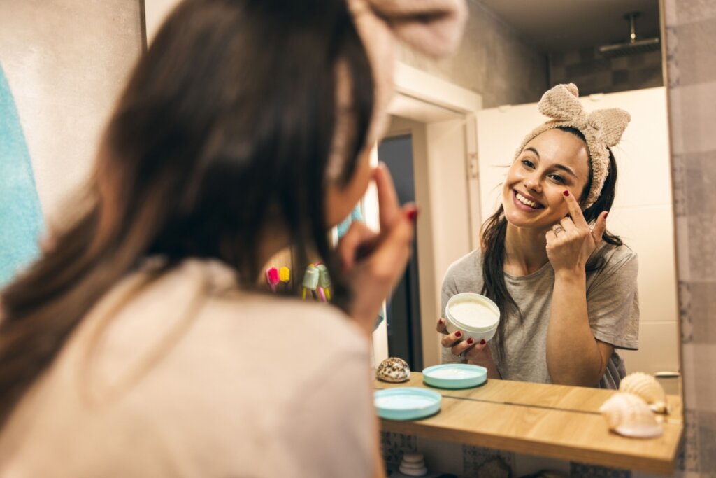 Mulher sorrindo, parada em frente a um espelho, e passando creme no rosto 