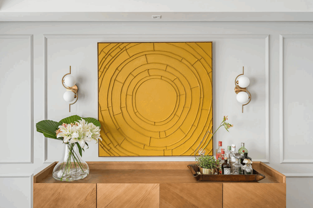 Parede branca com boiserie branca ao redor de quadro amarelo com círculos e com móvel em madeira encostado na parede e com luminárias de cada lado do quadro e artigos de decoração em cima do móvel 