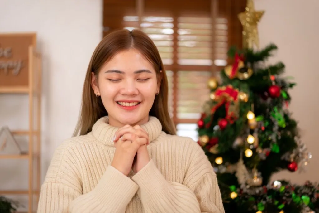 Jovem asiática com o cabelo solto, usando suéter bege rezando com árvores de Natal ao fundo