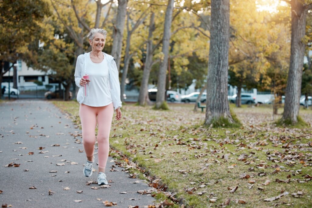 Mulher com cabelo curto, grisalho, usando camiseta e casaco branco e calça rosa, caminhando em parque, com uma garrafa de água na mão