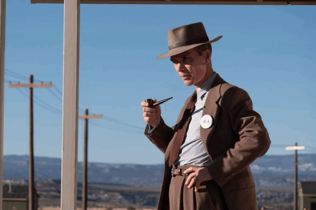 Homem com chapéu e paletó marrom segurando um cachimbo sob um céu claro em cena do filme Oppenheimer