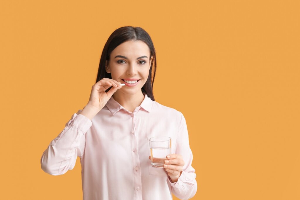 Mulher jovem de camisa rosa-claro segurando um copo de água e um comprimido, prestes a tomar a medicação, com fundo laranja