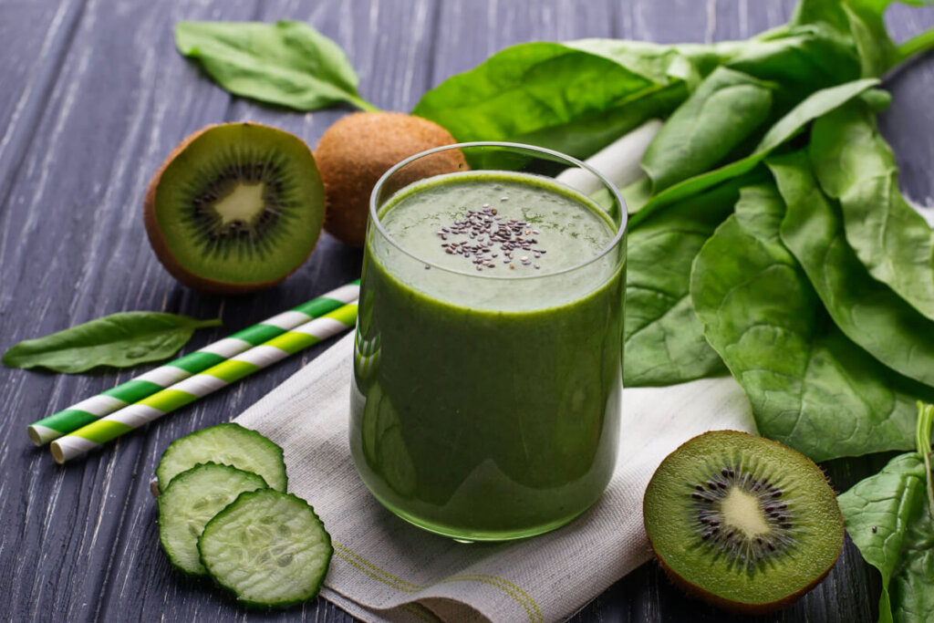 Suco de kiwi com couve e chia servido em copo de vidro com kiwi inteiro e um ao meio, com fatias de pepino e folhas de espinafre ao lado em cima de mesa de madeira e com canudo listrado em branco e verde