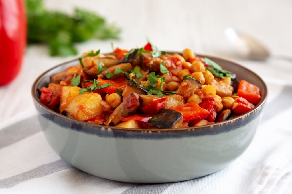 Prato com ensopado de berinjela, batata, tomate e grão-de-bico servido em tigela sobre tecido claro