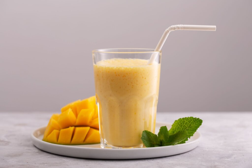 Copo de vidro com vitamina de manga e canudo branco. o copo está em prato branco com manga cortada e folhas de hortelã
