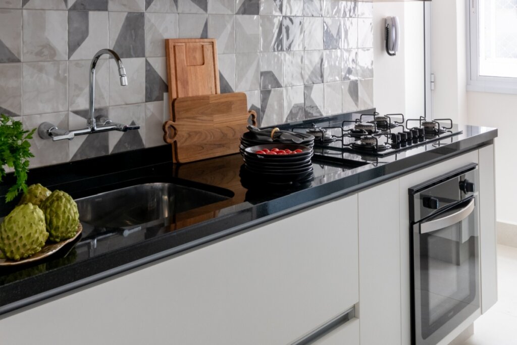 Close em uma bancada de granito preto com cuba de inox embutida. À esquerda, frutas verdes e um ramo de ervas; ao centro, pratos empilhados com tomates cereja e tábuas de corte de madeira encostadas no revestimento geométrico cinza da parede. À direita, um cooktop preto e um forno embutido em armários brancos.