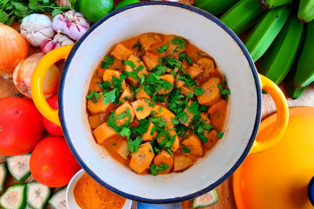Panela com moqueca de banana verde em molho alaranjado, finalizada com coentro fresco, cercada por tomates, alho e bananas