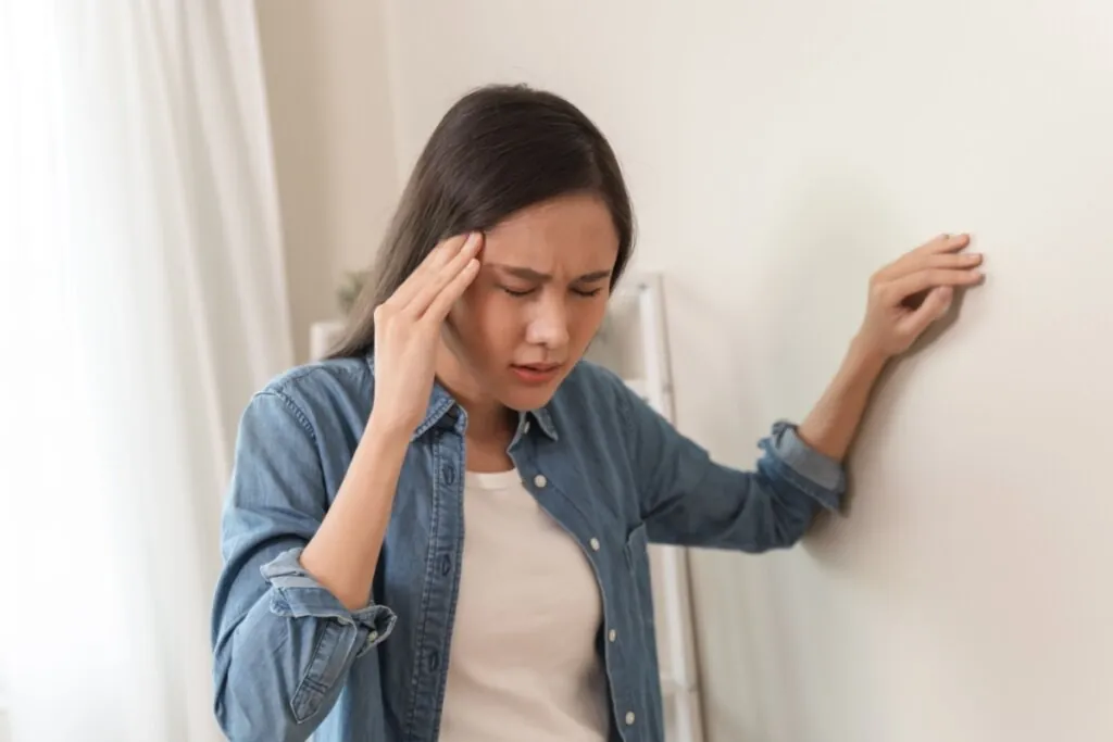 Uma mulher jovem, de cabelos escuros e lisos, veste uma camisa jeans sobre uma camiseta branca. Ela está com os olhos fechados e uma expressão de desconforto ou dor, pressionando os dedos de uma das mãos contra a têmpora, enquanto usa a outra mão para se apoiar em uma parede branca, sugerindo que sente tontura, mal-estar ou dor de cabeça.