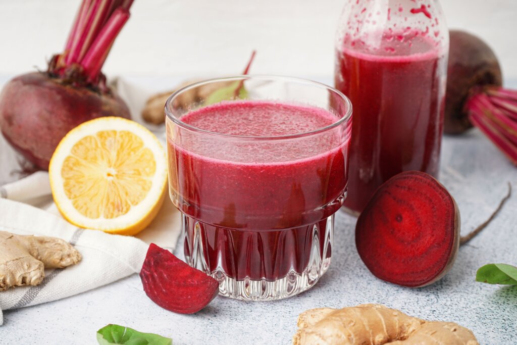 Um copo de vidro cheio de suco vermelho antioxidante de cor roxo (beterraba) e textura espessa. Ao redor do copo, estão ingredientes frescos: fatias e unidades inteiras de beterraba, gengibre e meio limão siciliano sobre um pano de prato claro, sugerindo uma bebida natural e saudável.