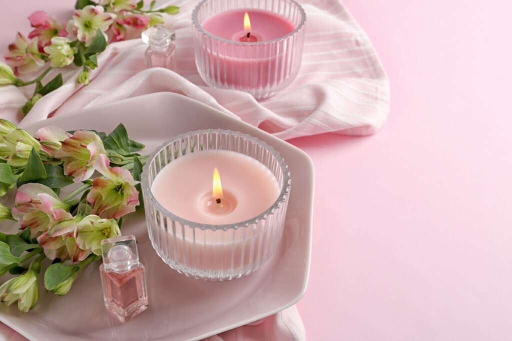 Duas velas rosas acesas em potes de vidro canelado sobre um prato e tecido rosa. A cena inclui flores alstroemérias e pequenos frascos de perfume, criando uma atmosfera delicada e relaxante.