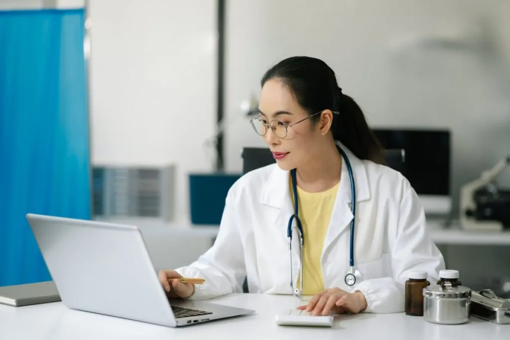 Médica de jaleco utilizando notebook e calculadora em consultório