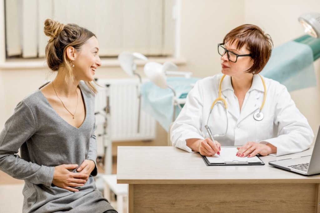 Mulher e médica sentadas em um consultório conversando; a paciente coloca a mão na barriga