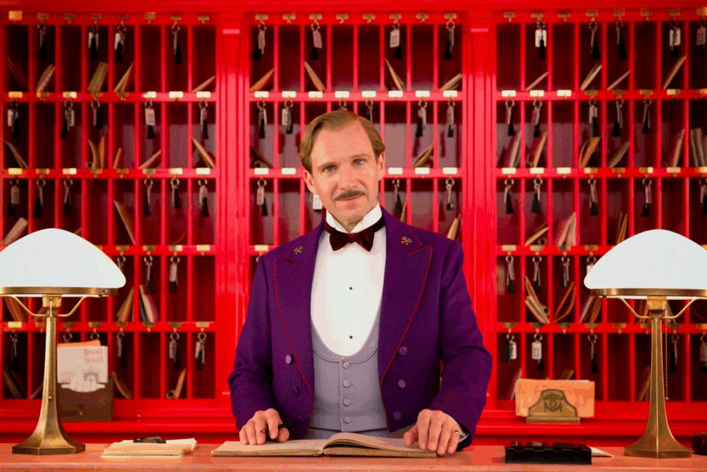 Homem com cabelo claro, curto, usando bigode e uniforme de hotel com camisa branca, gravata borboleta preta e colete e terno roxo, com parede de guarda volume vermelha ao fundo