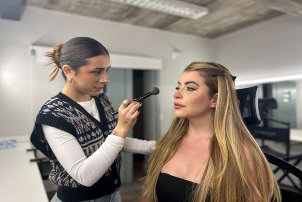 Maquiadora aplicando blush com pincel no rosto de uma cliente sentada em cadeira de estúdio