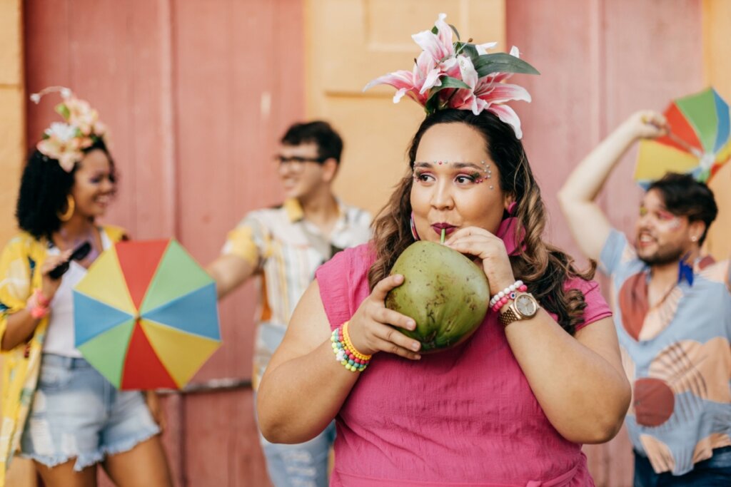 Mulher de camiseta rosa e adereços de carnaval tomando água de coco; pessoas fantasiadas para o Carnaval ao fundo