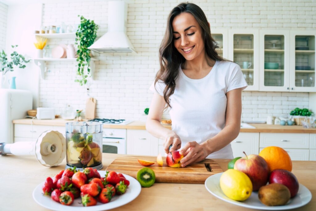 Mulher com cabelo longo, solto, usando camiseta branca cortando frutas em bancada para fazer suco