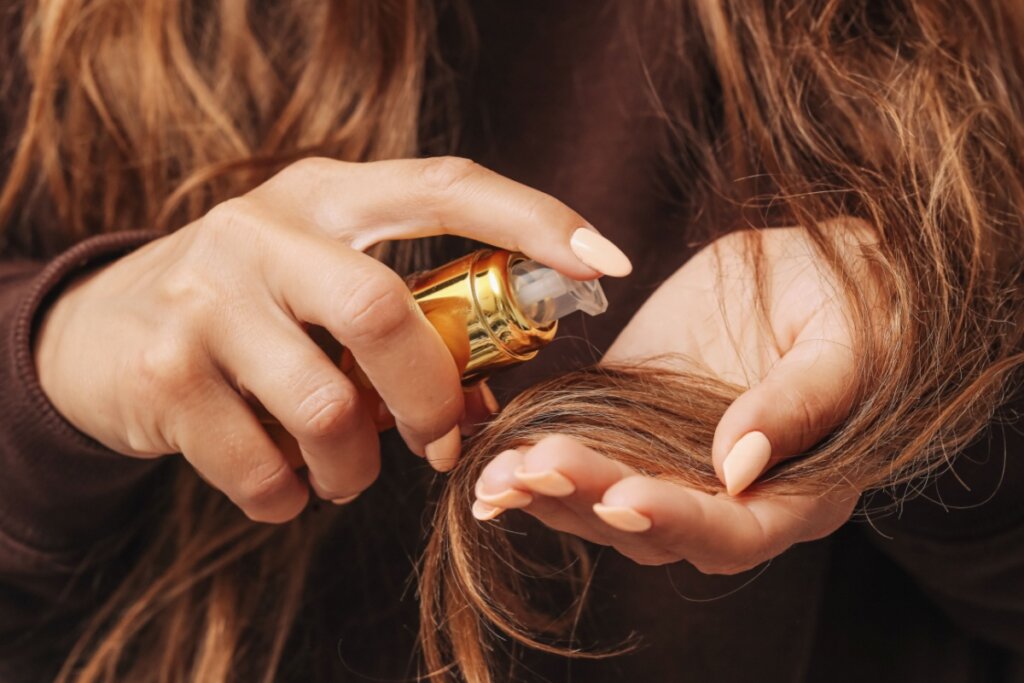 Mãos borrifando óleo no cabelo com um frasco transparente e dourado