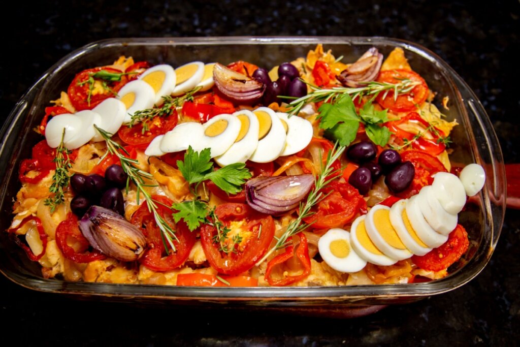 Vista superior de uma bacalhoada colorida e farta em um refratário de vidro retangular. O prato apresenta camadas de bacalhau, rodelas de tomates vermelhos maduros, pimentões, e pétalas de cebola roxa grelhada. Três fileiras de ovos cozidos fatiados atravessam o refratário. A finalização inclui azeitonas pretas pequenas, ramos de alecrim, tomilho e folhas de salsa fresca. O fundo é uma superfície escura de granito.