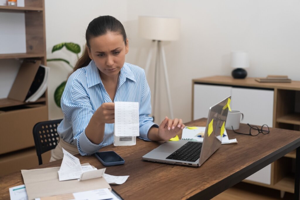 Uma mulher de camisa listrada azul está sentada à mesa em um escritório doméstico, com expressão de concentração e preocupação. Ela segura e analisa um longo comprovante de papel (recibo), enquanto a outra mão repousa sobre a mesa perto de um notebook aberto. Sobre a mesa de madeira, há diversos documentos, um smartphone, óculos e uma caneca. O notebook possui alguns adesivos amarelos (post-its) colados na tampa.