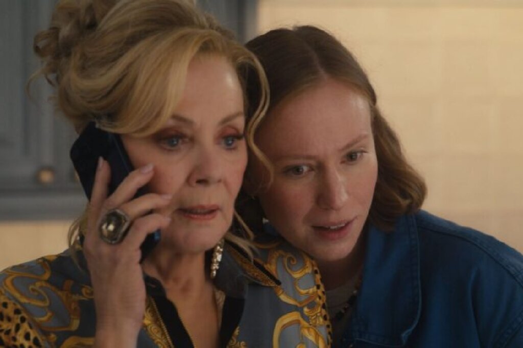 Close-up de duas mulheres. À esquerda, uma mulher mais velha com cabelos loiros presos em um penteado elegante, usando um brinco grande e um anel de destaque, fala ao celular com uma expressão preocupada. Ao lado dela, uma mulher mais jovem com cabelos castanhos claros observa a conversa com atenção e uma expressão de curiosidade, quase encostando o rosto no da outra.
