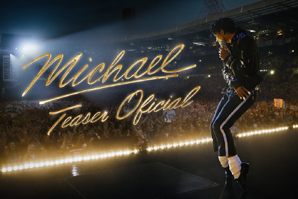 Imagem promocional da cinebiografia de Michael Jackson. Um ator caracterizado como o cantor está de perfil no palco de um estádio lotado, segurando um microfone. Ele usa uma jaqueta preta, calças com listras laterais brancas, meias brancas e sapatos pretos. No centro da imagem, o título "Michael Teaser Oficial" aparece escrito em letras douradas cursivas e brilhantes.