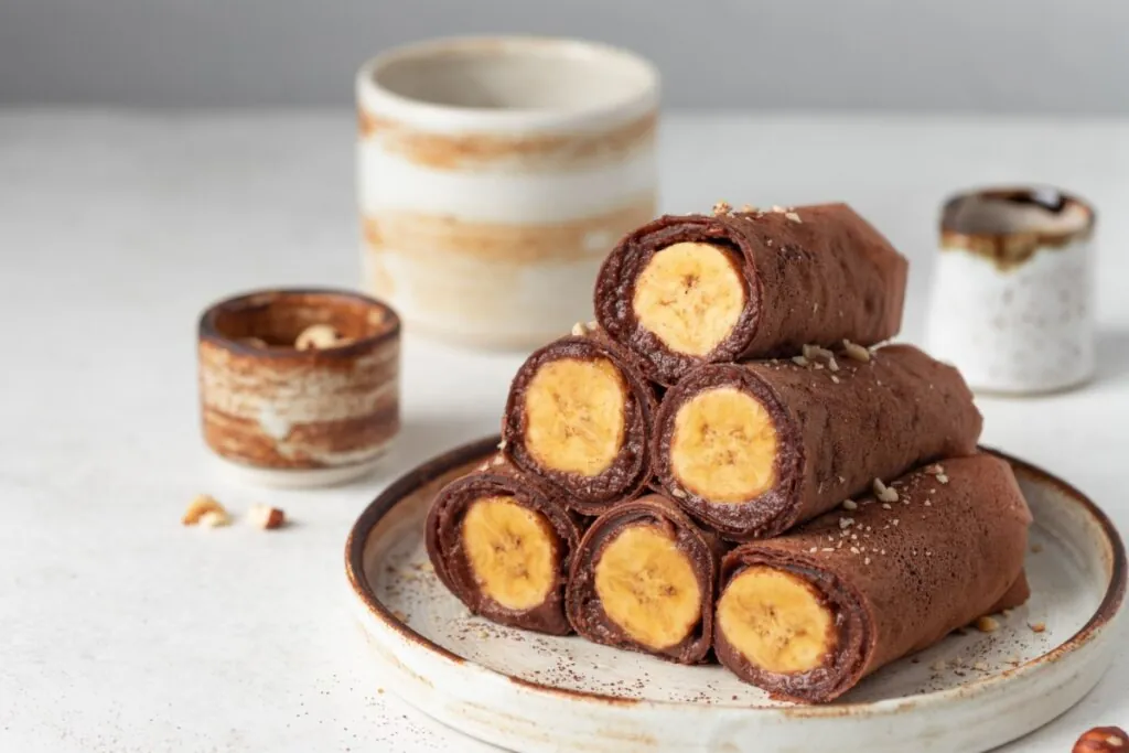 Rolinhos de massa de cacau recheados com banana, empilhados em prato com leve finalização de castanhas