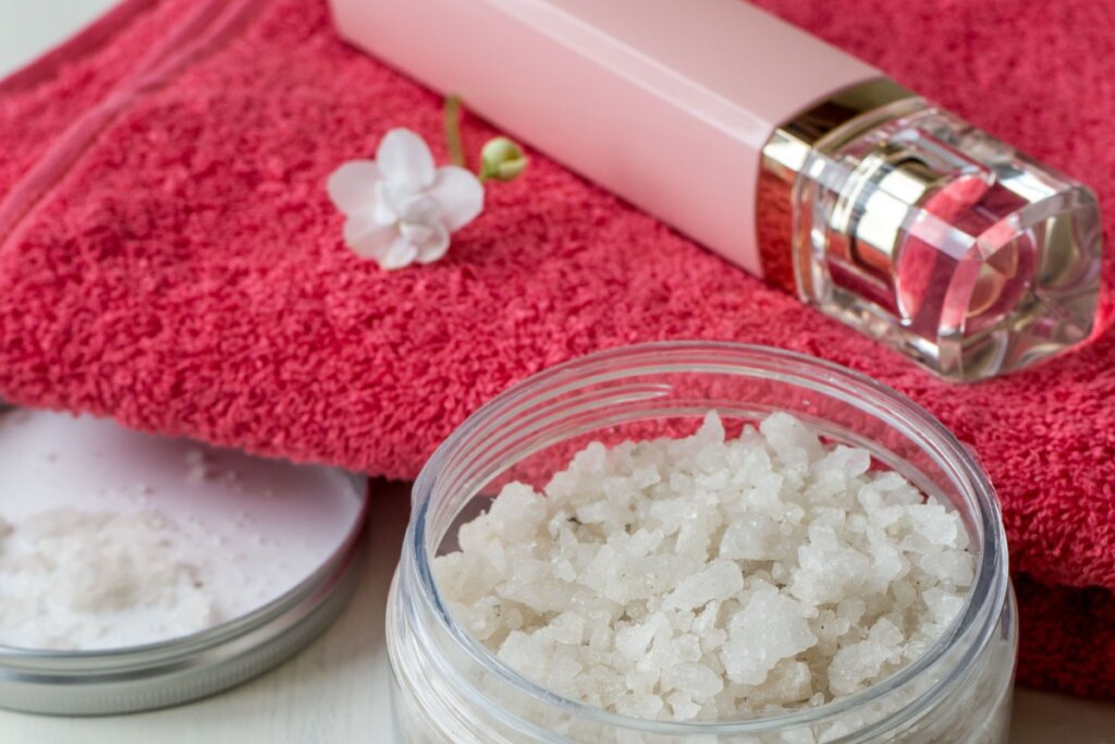 Close em um pote de vidro aberto contendo sal grosso em cristais brancos. Ao lado, a tampa metálica repousa sobre uma superfície clara. Ao fundo, uma toalha felpuda rosa vibrante serve de base para um frasco de perfume rosa com detalhes dourados e uma pequena flor branca decorativa.