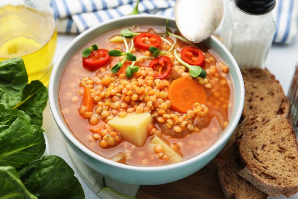 Tigela de sopa de lentilha com legumes, pimenta dedo-de-moça e pão rústico ao lado em mesa clara