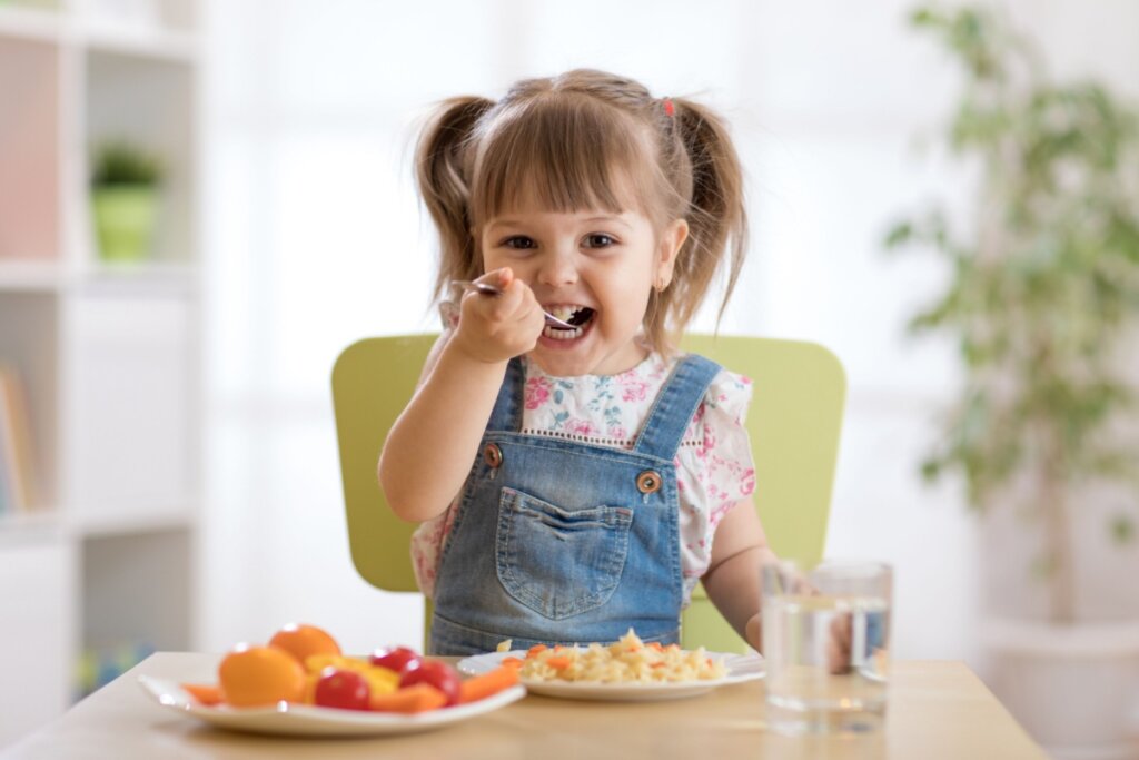 menina de jardineira azul sentada em mesa e levando colher com comida à boca. na mesa há prato com comida e copo com água