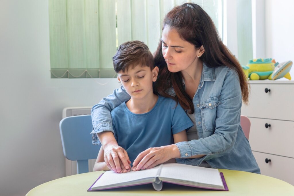Uma mulher sentada ao lado de um menino, os dois com as mãos sobre um livro de Braille aberto em uma mesa. A mulher veste uma jaqueta jeans e tem o braço esquerdo em volta dos ombros do menino. O menino veste uma camiseta azul e tem os olhos fechados. Seus dedos estão sobre as páginas em relevo do livro de Braille, com as pontas tocando os pontos. O livro é roxo com páginas brancas e espirais no meio. A mesa é redonda com tampo verde claro. Atrás deles, há uma cadeira azul claro e um móvel de gavetas branco. O fundo é iluminado por luz natural de uma janela com cortinas verticais verde claro.