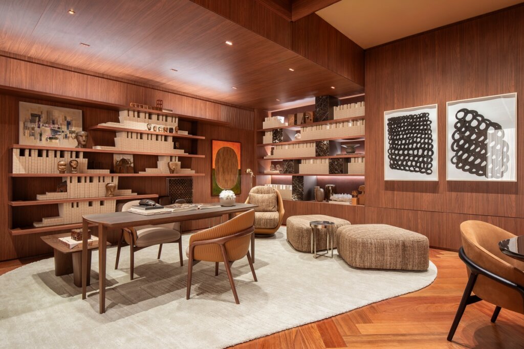 Vista de um escritório ou biblioteca sofisticada totalmente revestida em painéis de madeira de tom médio. Uma estante linear ocupa a parede esquerda, exibindo esculturas e livros de capas claras. No centro, uma mesa de trabalho elegante sobre um tapete circular off-white, acompanhada por poltronas de design moderno em tons de caramelo e marrom. A iluminação embutida no teto destaca as texturas amadeiradas.