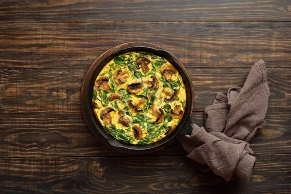 Uma fotografia de cima para baixo de um omelete em uma frigideira de ferro fundido, que repousa sobre uma tábua de madeira redonda. O omelete está coberta com cogumelos fatiados grelhados e folhas de espinafre frescas misturadas na mistura de ovos dourada. O cabo da frigideira aponta para a direita e está envolvido por um pano de cozinha de linho marrom-escuro amassado. O fundo é uma mesa de madeira de tábuas escuras com veios visíveis. A iluminação é natural e difusa.