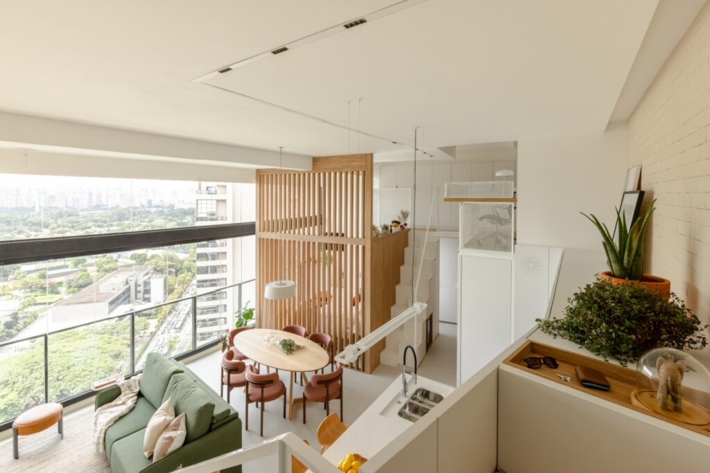 Vista aérea de um apartamento duplex com conceito aberto e decoração contemporânea. No nível inferior, destaca-se a sala de estar com sofá verde musgo e a mesa de jantar oval com cadeiras terracota. O ambiente é integrado a uma cozinha com bancada branca e pia de inox. Grandes janelas de vidro do chão ao teto oferecem uma vista panorâmica da cidade e áreas verdes. Uma estrutura de ripas de madeira centraliza o espaço, ocultando parcialmente a escada branca que leva ao mezanino. O uso de madeira clara, tons neutros e plantas cria uma atmosfera acolhedora e minimalista.
