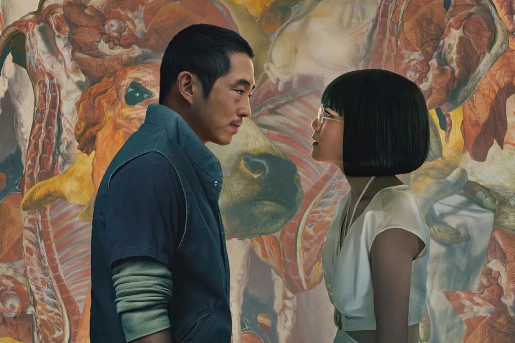 Imagem promocional da série "Beef" (Treta). Steven Yeun e Ali Wong estão de perfil, encarando-se fixamente com expressões de tensão e confronto. O fundo é uma pintura abstrata e expressiva que mistura tons de vermelho, bege e laranja, assemelhando-se a texturas de carne crua ou formas orgânicas intensas.