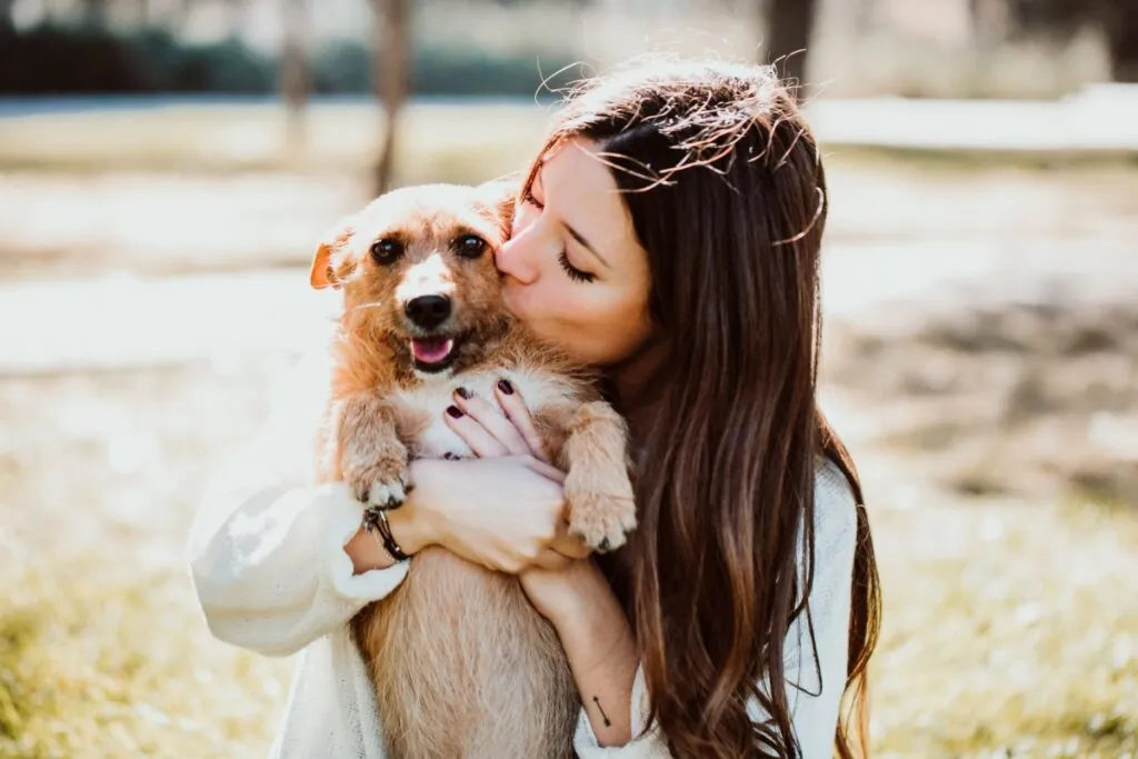 Mulher com cabelo solto, usando camisa de manga longa branca abraçada e beijando cachorro com pelagem bege ao ar livre