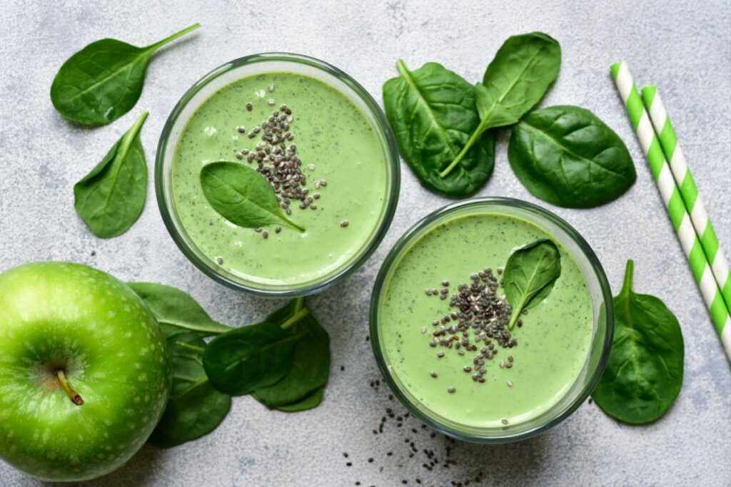 A imagem mostra dois copos de vitamina verde, com textura cremosa, preparados possivelmente com espinafre e maçã. Ao redor, aparecem folhas frescas de espinafre, uma maçã verde e sementes de chia