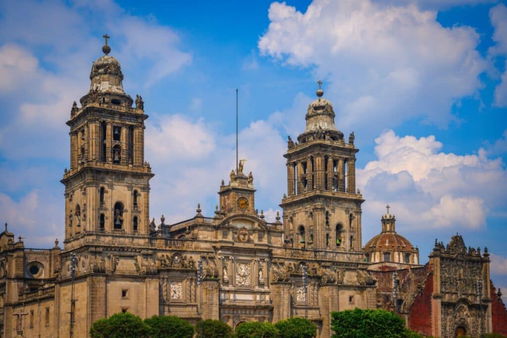 Uma vista frontal detalhada da Catedral Metropolitana da Cidade do México, mostrando as suas duas torres sineiras ornamentadas e a cúpula principal.