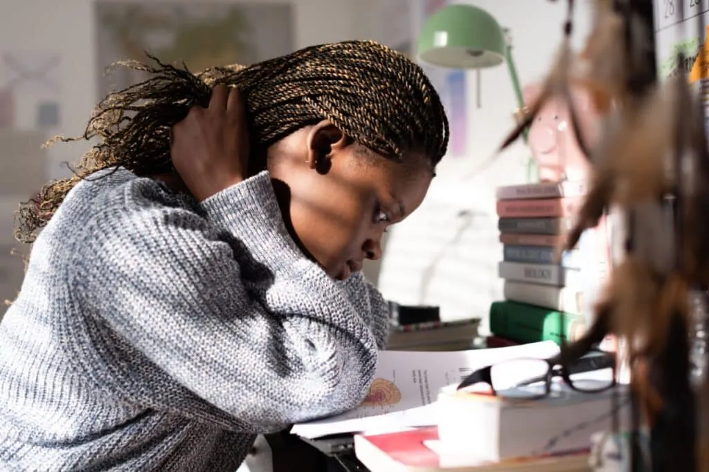 Uma jovem estudante negra, com tranças estilo box braids loiras e castanhas, concentrada na leitura de um livro didático aberto. Ela está sentada à sua mesa, inclinada para a frente, com a mão direita tocando o cabelo na nuca, pensativa. A mesa está repleta de materiais de estudo, incluindo uma pilha de livros coloridos, óculos de leitura sobre uma capa vermelha, uma luminária de mesa verde clara e um cofrinho rosa em forma de porco.