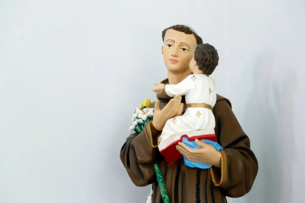 Estátua religiosa de Santo Antônio de Pádua contra um fundo branco liso. A escultura mostra o santo da cintura para cima, vestindo um hábito franciscano marrom com capuz e um cordão na cintura. Ele tem cabelos curtos e escuros com a tonsura (o topo da cabeça raspado). Santo Antônio segura carinhosamente o Menino Jesus em seus braços, que está sentado sobre uma Bíblia aberta com capa vermelha. O Menino Jesus veste uma túnica branca com detalhes dourados e está de costas, olhando para o rosto de Santo Antônio. No braço direito do santo, há também um ramo de lírios brancos.
