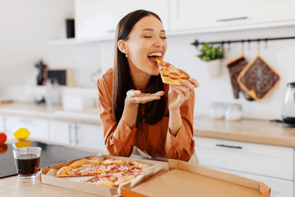 Mulher jovem e sorridente em uma cozinha, prestes a dar uma mordida em uma fatia de pizza de calabresa. À sua frente, na bancada, há uma caixa aberta com o restante da pizza e um copo com refrigerante.