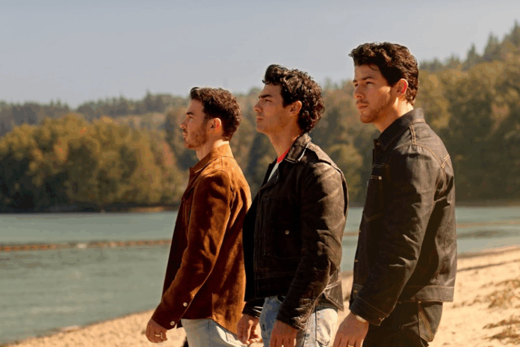 Fotografia dos três integrantes da banda Jonas Brothers (Kevin, Joe e Nick Jonas) alinhados de perfil em uma praia com vegetação ao fundo, olhando para a esquerda com expressões sérias. Eles vestem jaquetas casuais — uma marrom de camurça, uma preta de couro e uma de jeans escuro. A luz do sol ilumina a cena de forma calorosa e dourada.