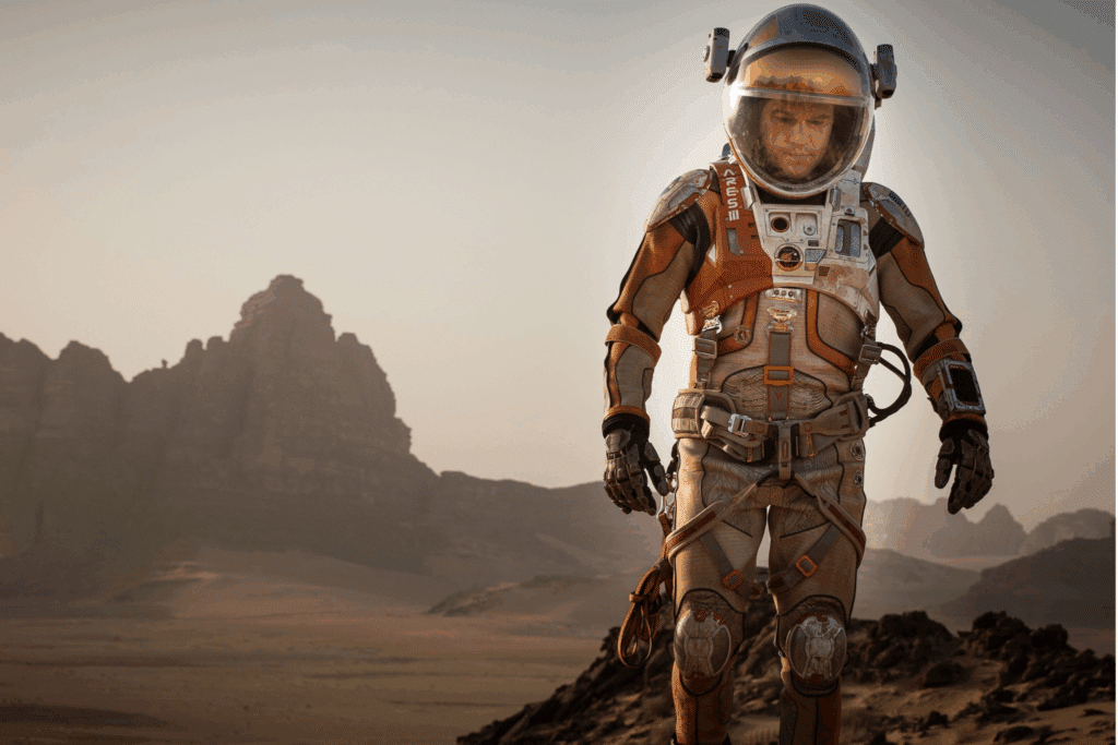 Homem vestindo um traje espacial completo nas cores branco e laranja, caminhando por um terreno desértico e rochoso que remete ao planeta Marte, sob um céu de tom claro.